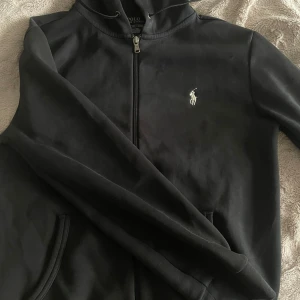 Svart hoodiejacka från Polo Ralph Lauren - Svart hoodiejacka från Polo Ralph Lauren med dragkedja framtill och klassisk broderad logga på bröstet. Jackan har huva med snörning och två fickor. Tillverkad i mjuk bomull, perfekt för en chill och stilren look. Den är i xs men passar s