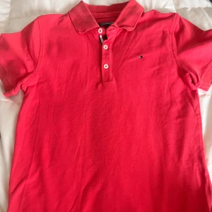 Rosa tajt pikétröja från Tommy Hilfiger - Snygg rosa tajt pikétröja från Tommy Hilfiger med klassisk krage och tre knappar framtill. Tröjan har korta ärmar och det ikoniska Hilfiger-loggan broderad på bröstet. 