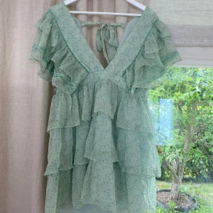 Mintgrön volangklänning med blommor - Supersöt mintgrön kortklänning med småblommigt mönster, volanger i flera lager och djup v-ringning med knyt i nacken. Klänningen är i ett tunt, lätt och transparent material som känns perfekt för varma dagar.