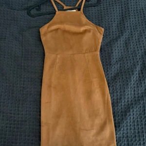 Brun mockaklänning med smala axelband - Snygg brun klänning i mockaimitation med smala axelband och figurnära passform. Klänningen har en enkel, stilren design utan mönster och är perfekt för dig som gillar en minimalistisk look. Materialet känns mjukt och ger en cool vintagekänsla. Klänningen har mindre slitage och några trådar synliga i sömmen baktill (se bild). 