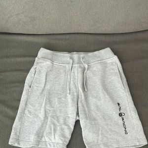 Gråa shorts från Sail Racing - Snygga ljusgrå shorts från Sail Racing med bekväm resår och snörning i midjan. Mjukt bomullsmaterial och två sidofickor. Logga tryckt längs ena benet för en sportig vibe. Perfekta för sommardagar eller chill hemma. Stolek 160 cm