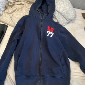 Mörkblå hoodie Sail Racing SR77 - Mörkblå hoodie från Sail Racing med dragkedja och huva. Broderad SR i rött och 77 i vitt på bröstet. Mjuk insida och praktiska fickor. Perfekt för dig som gillar sportig och avslappnad stil.