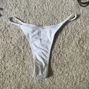 Vit bikiniunderdel med smala band - Säljer en vit bikiniunderdel med smala elastiska band i sidorna och hög skärning. Enkel och stilren design som ger en clean look på stranden eller vid poolen. Materialet är stretchigt och bekvämt, perfekt för sommardagar.