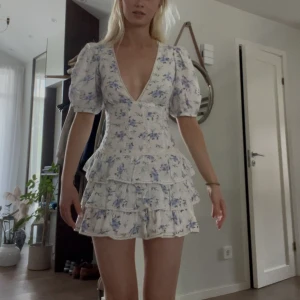 Blommig volangklänning från Zara XS - Supersöt kort klänning från Zara med puffärmar, djup v-ringning och flera volanger. Vit botten med blått och lila blommönster. Tillverkad i bomull, perfekt för sommarens alla tillfällen. Sitter tajt upptill och har en flowy kjol.