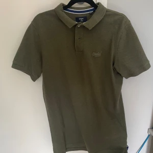 Olivgrön pikétröja från Superdry - Säljer en olivgrön pikétröja från Superdry i klassisk modell med krage och knappar. Tröjan har korta ärmar och diskret broderad logga på bröstet. Kragen har en snygg vit och marinblå randig detalj. Perfekt för en clean och avslappnad stil.