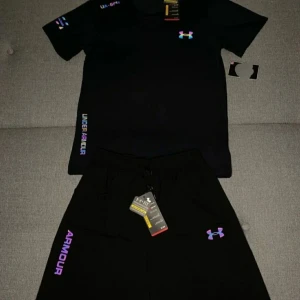 Under armour set med reflex - Svart t-shirt från Under Armour med korta ärmar och färgglad regnbågsskimrande logga på bröstet. Snygga detaljer med tryck på ärmen och sidan. Perfekt för dig som gillar sportig och modern stil. Materialet är lätt och andas, vilket gör den grym för träning. Priset är ej hugget i sten !