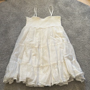 Vit somrig kortklänning med volang - Luftig vit kortklänning med smock upptill och tunna axelband. Klänningen har flera volanglager och är tillverkad i ett lätt, skrynkligt tyg som ger en avslappnad känsla. Perfekt för varma dagar och enkel att styla.