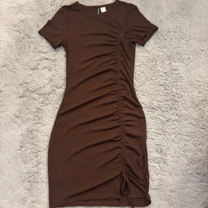 Brun kort klänning med snörning - Snygg brun kortklänning från Bershka med tight passform och korta ärmar. Klänningen har en cool snörningsdetalj längs sidan som ger en rynkad effekt och gör looken extra trendig. Perfekt för dig som gillar stilrena och moderna plagg.