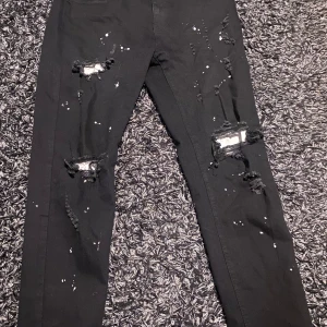 Svarta skinny jeans med slitningar - Svarta skinny jeans med stora slitningar och hål på knäna samt vita färgstänk över hela byxorna. Jeansen har klassisk femficksdesign och smal passform. Perfekta för dig som gillar en edgy och trendig streetstil.