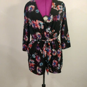 Svart blommig kort jumpsuit med bälte - Kort svart jumpsuit med färgglatt blommönster i rött, blått, lila och vitt. Omlottmodell med v-ringning, trekvartsärm och knytbälte i midjan. Perfekt för dig som gillar färg och vill sticka ut.