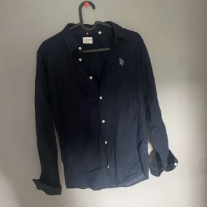 Mörkblå skjorta från US Polo Assn - Snygg mörkblå skjorta från US Polo Assn med klassisk krage och vita knappar. Skjortan har lång ärm och broderad logga på bröstet. Perfekt för dig som gillar stilrena plagg med en sportig touch.