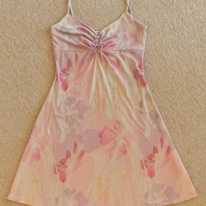 Pastellblommig kort klänning - Superfin kort klänning med blommigt mönster i pastellrosa, lila och beige toner. Smala axelband, v-ringning med snygg snörningsdetalj framtill. Perfekt för varma dagar och sommarvibbar.