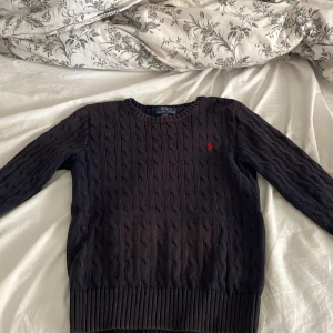 Mörkblå kabelstickad tröja Polo Ralph Lauren - Tröja från Ralph lauren i storlek 10-12y. Den är väldigt skön. Tyvärr är det som ett väldigt litet hål som ni ser på bild 3. Tröjan är lite solblekt men ändå inget som man tänker på.