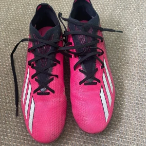 Rosa Adidas fotbollsskor med svarta detaljer.   - Snygga fotbollsskor från Adidas i en stark rosa färg med svarta och vita detaljer. Skorna har klassiska tre ränder på sidan, svart snörning och svart häl med X-logga. Ovandelen är i syntetmaterial och designen är smal och sportig. 