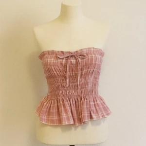 Rutig rosa bandeau tubtopp med volang - Supersöt bandeau tubtopp i rosa med rutigt mönster och volang nertill. Smockad överdel och söt knytning framtill ger en romantisk vibe. Perfekt för varma dagar och lätt att matcha med jeans eller kjol.