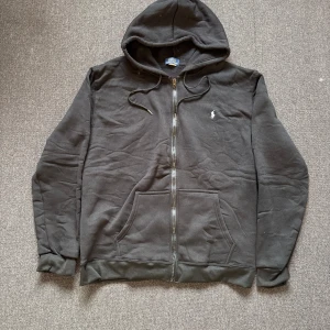 Svart set från Polo Ralph Lauren - Svart hoodie från Polo Ralph Lauren med dragkedja, huva med snörning och klassisk vit broderad logga på bröstet. Mjuk bomullskvalitet och känguruficka framtill. Perfekt för chill dagar eller när du vill ha en clean streetstyle.