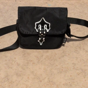 Svart axelväska från Trapstar - Säljer en svart axelväska från Trapstar med justerbart band och unik vit-svart spänndetalj framtill. Väskan har meshstruktur på framsidan och cool logga på sidan. Perfekt för att bära det viktigaste och ge din outfit en streetwear vibe.