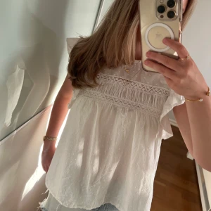 Vit blus med spets från Vero Moda - Supersöt vit blus från Vero Moda med volangärmar och broderade spetsdetaljer över bröstet. Blusen är ärmlös med lätt rynk och har en romantisk känsla. Perfekt till jeansshorts eller kjol för en fräsch sommarlook.