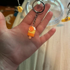Nyckelring med glass i orange och vitt - Söt nyckelring med en miniatyrglass i orange, vitt och gult. Glassen har en liten apelsinskiva på toppen och hänger i en silverfärgad kedja. Perfekt accessoar för att piffa upp dina nycklar eller väska.