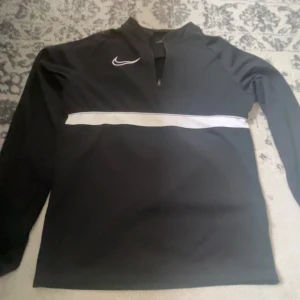 Svart Nike half zip tröja - Svart långärmad tröja från Nike med vit logga på bröstet och en vit horisontell rand över magen. Tröjan har half zip framtill och är gjord i ett mjukt, sportigt material som passar perfekt till träning eller chill.