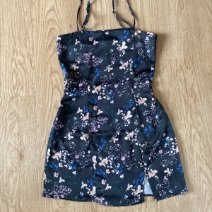Blommig kort klänning från Pull&Bear storlek S - Supersöt kort klänning från Pull&Bear med smala axelband och svart botten. Storlek S men tycker att den passar mer som XS. Klänningen har ett blommigt mönster i rosa, blått och beige, samt en liten slits framtill. Sval och lätt, perfekt för varma dagar eller när du vill känna dig extra fin.                                   OBS! Klänningen är justerad i midjan för att få en tajtare passform som syns på bild 4 & 5. Detta går att sprätta upp om så behövs. 