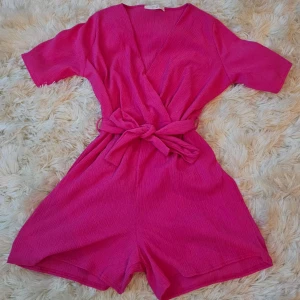 Rosa omlottjumpsuit Sisters Point XL - Superfin kort rosa omlott jumpsuit från Sisters Point i storlek XL. Jumpsuiten har knytband i midjan, v-ringning och korta ärmar. Materialet är lätt och stretchigt i 95% polyester och 5% elastan, perfekt för varma dagar.
