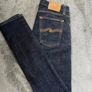 Mörkblå jeans från Nudie Jeans - Snygga mörkblå jeans från Nudie Jeans med klassisk gul kontrastsöm och läderpatch bak. Modellen har raka ben och normal passform, tillverkade i slitstarkt jeanstyg. Perfekta för dig som gillar stilrena och tidlösa jeans. Kom gärna med bud.( BÄST PRISET NI KOMMER HITTA!!) srlk: 26/32