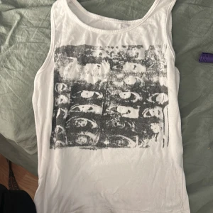 Vit linnetopp med svart tryck från SHEIN - Säljer en vit linnetopp från SHEIN i storlek XS. Toppen är ärmlös och har ett svart, konstnärligt tryck framtill med ansikten i rutmönster. Mjuk och lätt i materialet, perfekt för en chill och trendig look.
