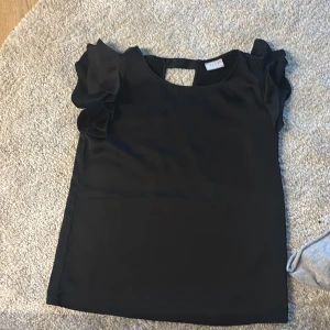 Svart blus med volangärmar från VILA - Supersnygg svart blus från VILA med volangiga korta ärmar och en djup v-ringning i ryggen. Toppens material är lätt och har en elegant känsla, perfekt när du vill känna dig lite extra. Enkel men ändå med coola detaljer som gör att den sticker ut.