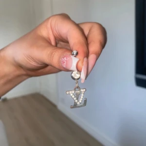 LV navelpiercing i silver med stenar - Snygg navelpiercing i silverfärg med hängande LV-logga dekorerad med glittrande stenar. Piercingen har en böjd stav och två kulor, varav den ena är prydd med en klar sten. Perfekt accessoar för dig som vill sticka ut lite extra.