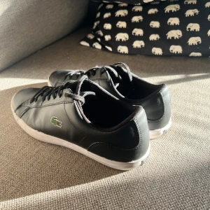 Svarta  Lacoste Skor - Stilrena svarta sneakers från Lacoste med klassisk krokodillogga på sidan. Skorna har snörning, vit platt sula och är tillverkade i skinn för en clean look. Storlek 44. Inprincip nyskick 