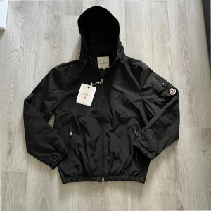 Svart vindjacka från Moncler med huva - Snygg svart vindjacka från Moncler med huva och dragkedja framtill. Jackan har två fickor med dragkedja och elastisk nederkant. Klassisk Moncler-logga på ärmen ger en exklusiv vibe. Perfekt för dig som vill ha en stilren och funktionell jacka. NFC fungerar 