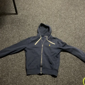 Mörkblå hoodie från Ralph Lauren - Säljer en snygg mörkblå hoodie från Ralph Lauren med dragkedja och gul logga på bröstet. Den har en justerbar huva med beige snören och ribbade muddar vid ärmslut och nederkant. Perfekt för en avslappnad stil.