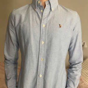 Ljusblå skjorta från Ralph Lauren - Snygg ljusblå skjorta från Ralph Lauren med klassisk krage och broderad logga på bröstet. Skjortan har långa ärmar och knappar framtill. Bra skick. Jag är 170 cm och väger lite mer än 50 kg