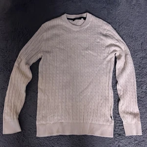 Beige stickad tröja från Jack & Jones - Säljer en stilren beige stickad tröja från Jack & Jones. Tröjan har ett klassiskt kabelstickat mönster och långa ärmar med ribbade muddar. Perfekt för kyligare dagar.
