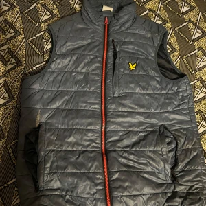 Grå quiltad väst med dragkedja - Snygg grå quiltad väst med röd dragkedja och en gul logga på bröstet. Västen har en hög krage och praktiska fickor framtill. Perfekt för lager-på-lager under kyligare dagar.