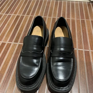 Svarta loafers från H&M 37 - Snygga svarta loafers från H&M i skinn med en robust sula. Perfekta för en stilren look med en klassisk design. Skorna har en dekorativ rem över vristen och en bekväm passform. Fint skick och sköna.