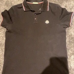 Svart pikétröja från Moncler - Säljer en stilren svart pikétröja från Moncler med klassisk krage och knappar framtill. Tröjan har korta ärmar med röd och vit randdetalj vid ärmslut och krage. Perfekt för en avslappnad men ändå snygg look.