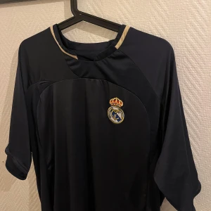Svart fotbollströja från Real Madrid - Snygg mörkblå fotbollströja Real Madrid. ✅Tröjan har korta ärmar och en stilren design med gulddetaljer vid kragen. Storlek L ✅ 110kr ✅