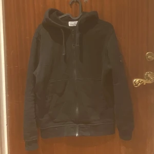Svart hoodie från Stone Island - Säljer en stilren svart hoodie från Stone Island med dragkedja och huva. Tröjan har en broderad logga på ärmen och är perfekt för en casual look. Den är tillverkad i ett mjukt material som ger en bekväm passform. Inga defekter hör av er vid funderingar 👍