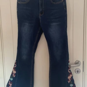 Blå bootcut jeans med blommigt broderi från Bamboo - Snygga blå bootcut jeans från Bamboo med vackert blommigt broderi längs benen. Perfekta för att ge en unik touch till din outfit. Högmidjade med klassisk femficksdesign och knappgylf. Säljs till högsta bud!❤️