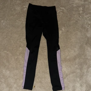 Svarta leggings från SOC - Snygga svarta leggings från SOC med lila detaljer längs sidorna. Perfekta för träning med ett tekniskt material som andas och håller dig torr och bekväm. Elastisk midja för en bra passform.