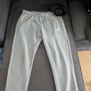 Grå mjukisbyxor från Nike - Säljer ett par grå mjukisbyxor från Nike med elastisk midja och snörning. Byxorna har en broderad Nike-logga på sidan och är perfekta för en avslappnad stil. De är tillverkade i mjukt material för extra komfort.