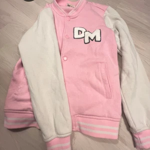 Rosa och vit hoodiejacka från WAI WAI - Säljer en söt rosa och vit jacka som har modellen som en jacka men material som en hoodie! från WAI WAI med DM-bokstäver på bröstet. Jackan har långa ärmar och stängs med knappar framtill. Perfekt för en cool och avslappnad stil.