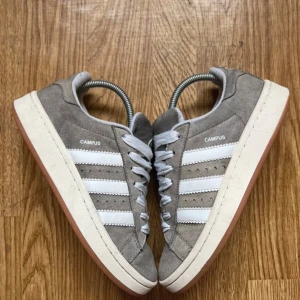 Grå Adidas Campus sneakers - Snygga grå Adidas Campus sneakers i mocka med vita detaljer och klassiska tre ränder på sidan. Skorna har en vit sula och brunt gummi undertill. Perfekta för en stilren look. Kan sänka priset vid snabb affär