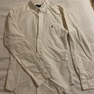 Vit skjorta från Ralph Lauren - Säljer en klassisk vit skjorta från Ralph Lauren med broderad logga på bröstet. Skjortan har långa ärmar och knappar framtill. Perfekt för en stilren look. Nypris 1400
