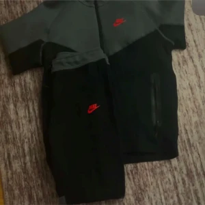 Grå och svart hoodie från Nike - Säljer en grå och svart hoodie från Nike med korta ärmar och dragkedja. Den har en röd Nike-logga på bröstet och två fickor framtill. Perfekt för en sportig look!