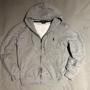 Grå zip hoodie  från Ralph Lauren - Säljer en stilren grå hoodie från  Ralph Lauren med dragkedja och det klassiska logotypbroderiet på bröstet i storlek M men skulle säga att den är mer storlek S. Tröjan har en bekväm passform och är perfekt för en avslappnad look. Den är tillverkad i mjukt material och har ribbade muddar vid ärmslut och nederkant. 