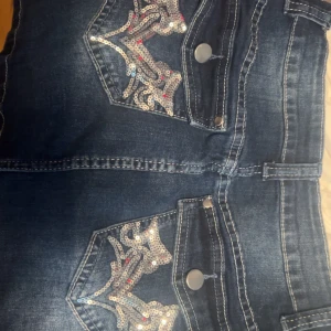 Blå jeanskjol med paljettdetaljer - Snygga blå jeans med paljetter på bakfickorna som ger en extra touch. De har en klassisk femficksdesign och stängs med dragkedja och knapp. Perfekta för att lägga till lite glitter i vardagen.