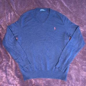 Blå tröja från Ralph Lauren - Säljer en stilren blå tröja från Ralph Lauren med v-ringning och det klassiska broderade logotypen i rött på bröstet. Tröjan har långa ärmar och ribbade muddar vid ärmslut och nederkant. Perfekt för en avslappnad men ändå elegant look.